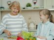 Senior grandmother is making salad and feeding cute happy child with vegetable chops expressing love having fun in kitchen at home. People and lifestyle concept.