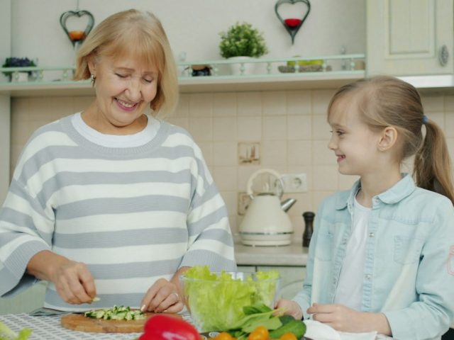Senior grandmother is making salad and feeding cute happy child with vegetable chops expressing love having fun in kitchen at home. People and lifestyle concept.